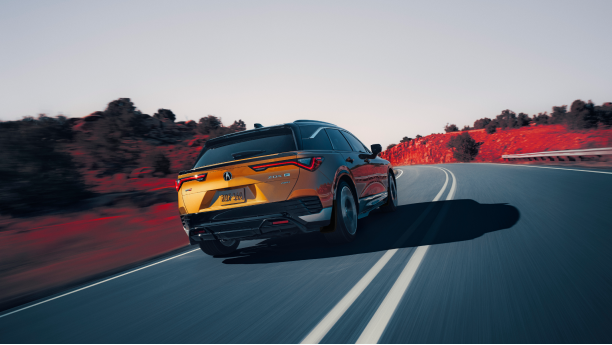 3/4 rear view of gold ZDX driving on mountain highway. Part of the image is saturated with colour, so the dirt on the sides of the highway is red. 