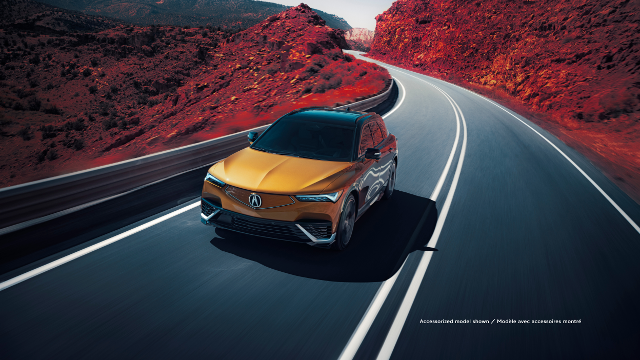 3/4 bird’s eye front view of gold ZDX driving on mountain highway. Part of the image is saturated with colour, so the dirt on the sides of the highway is red.