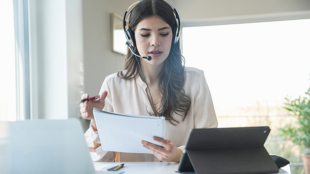 A young woman wearing a headset works in front of a laptop and tablet. / Une jeune femme portant un casque d’écoute travaille devant un ordinateur portable et une tablette.