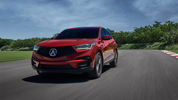 Front, slightly angled view of a red Acura RDX driving around a track surrounded by trees and grass. // Vue avant latérale d’un Acura RDX rouge roulant sur une piste entourée d’arbres et de gazon.