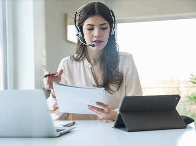 Image of a young woman with long brown hair, wearing a headset and appearing to speak into the microphone as she looks over paperwork at a desk./Image d'une jeune femme aux longs cheveux bruns, portant un casque d’écoute et semblant parler dans le microphone alors qu'elle regarde la paperasse sur un bureau.